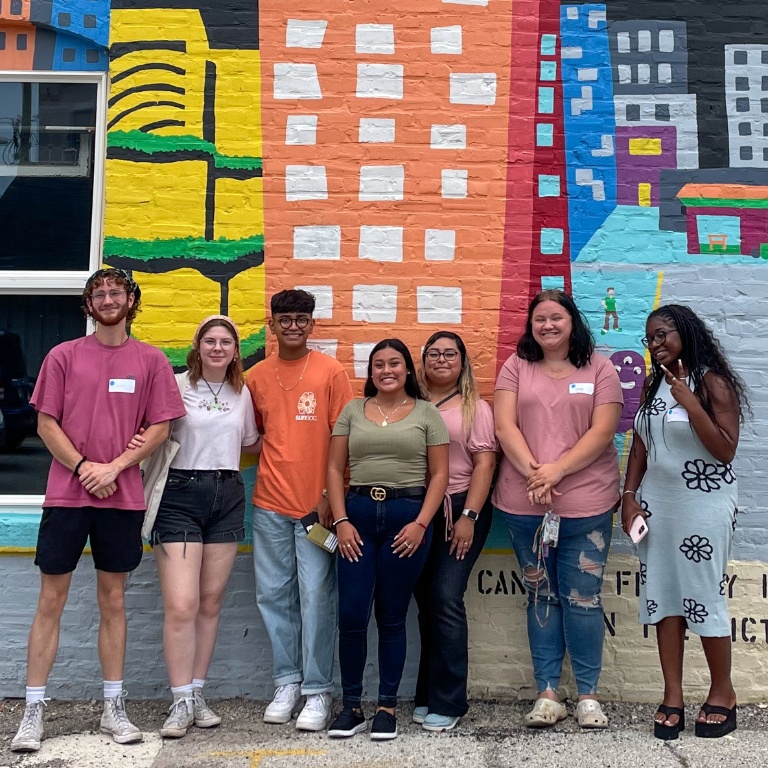 A group of students pose in front of a wall mural. 