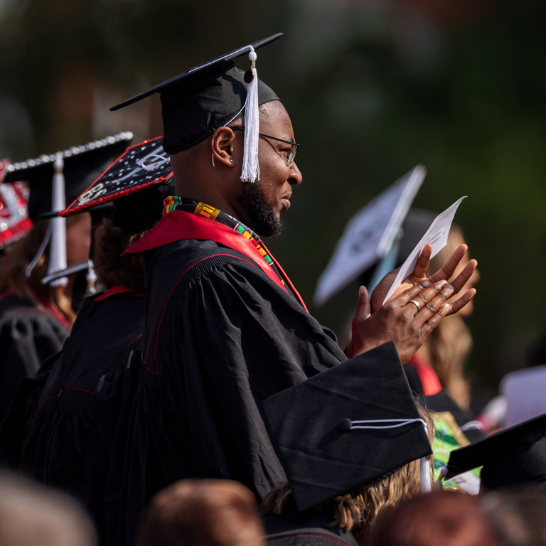 A bearded IUK graduate applauds in the middle of a crowd