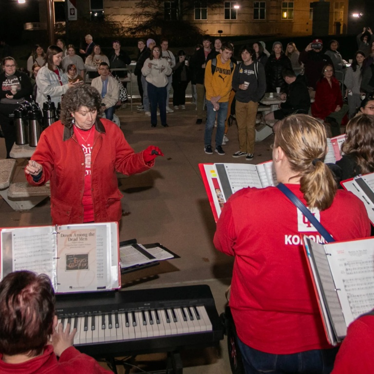 Conductor and choir performing outdoors with a crowd in the background.